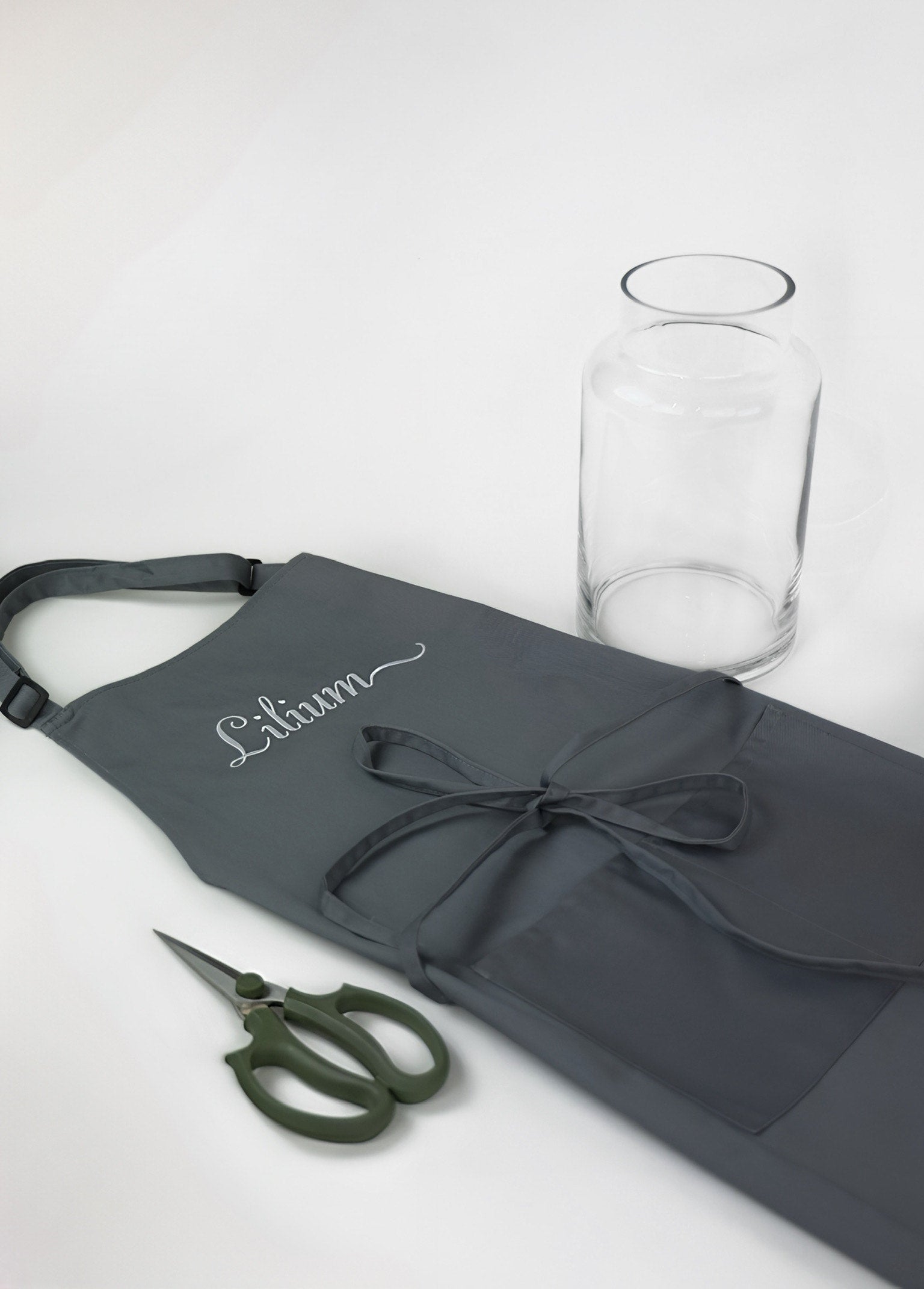 Gray apron with 'Lilium' embroidery, green scissors, and clear glass on a white background